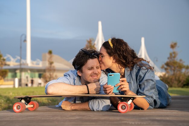 日没時に寒いスマートフォンを見て屋外のスケートボードに横たわって恋にスタイリッシュな若いカップル