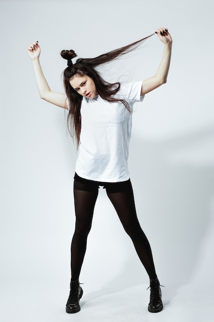 Photo stylish young dark-haired girl with a funny hairstyle dressed in white t-shirt, black tights and shoes is posing on the white background in the studio .