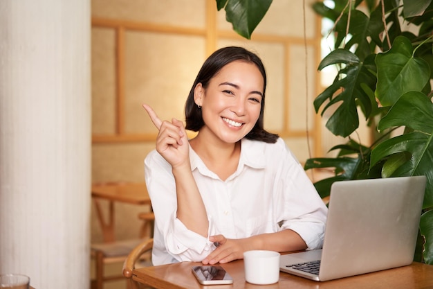 Stylish young entrepreneur girl with laptop sitting in cafe pointing finger at advertisement banner