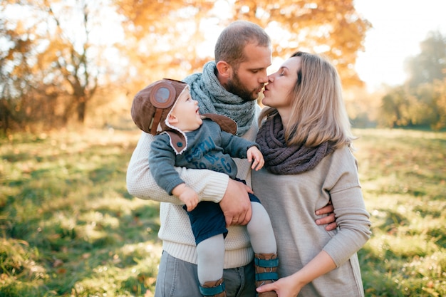 Stylish young family holding baby in hands and kissing in sunny park.