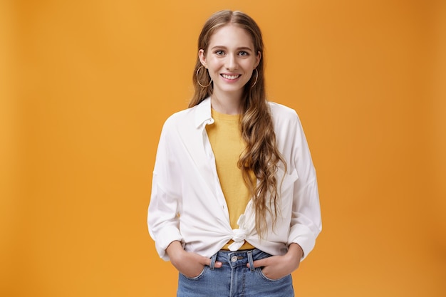 Stylish young female designer wants help friend shopping holding hand in pockets smiling joyfully and self-assured at camera wearing trendy blouse over yellow t-shirt posing over orange wall.