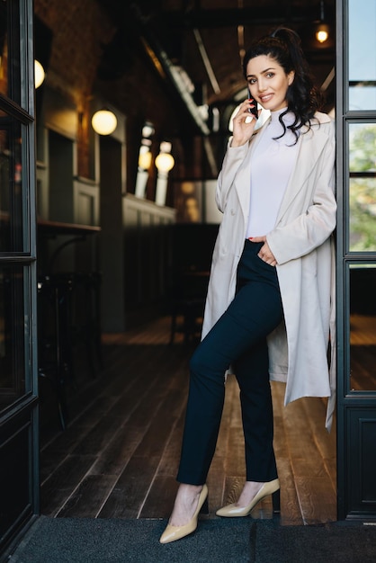 Stylish young female with dark hair wearing white coat black trousers and shoes with heels posing into camera while standing against cafe background chatting over phone holding hand in pocket