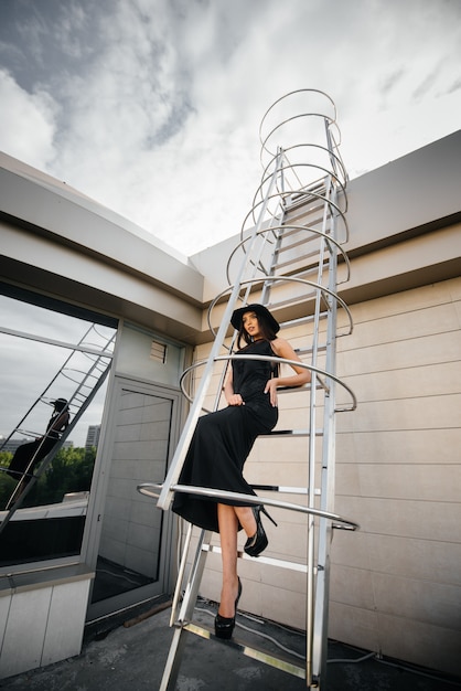 Stylish young girl posing in a hat on the fire escape of the business center. Fashion.