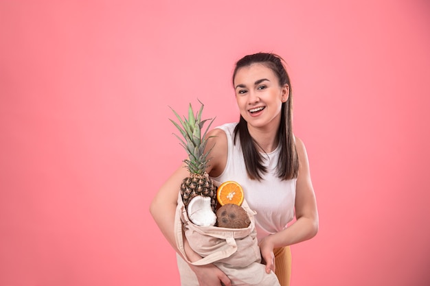 Stylish young girl smiling and holding an eco bag with exotic fruits on a pink wall copy space.