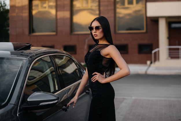 Stylish young girl stands near the car in a black dress. Business fashion and style.