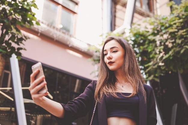 スタイリッシュな若い女の子は、selfieを取ります。女の子は、スマートフォンで彼女の写真を作ります。