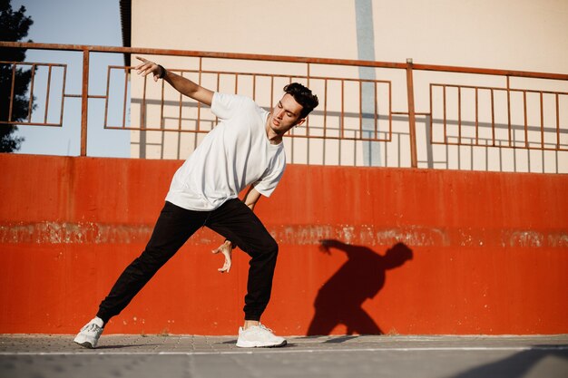 Stylish young guy dressed in jeans and white t-shirt is dancing brakedance in the street against a painted concrete wall in the sunny day .