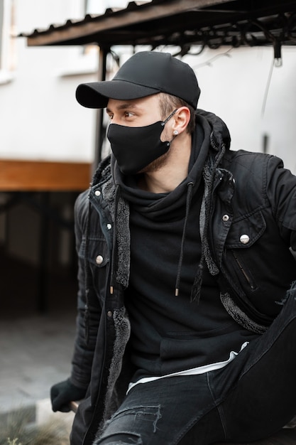 Stylish young guy with a black protective mask in fashionable black clothes with a jacket, a hoodie and a cap sits on the street. Urban male casual style and coronavirus concept