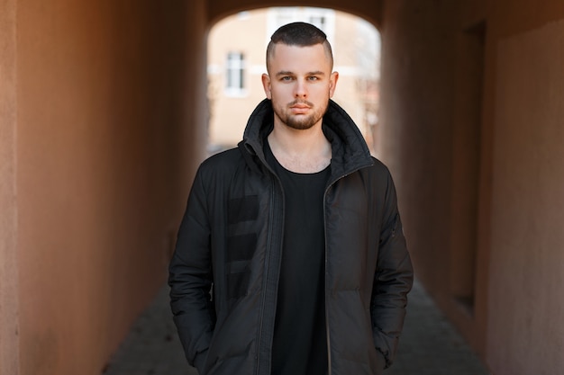 Stylish young handsome man with a short haircut in a black fashion winter jacket stands on the street