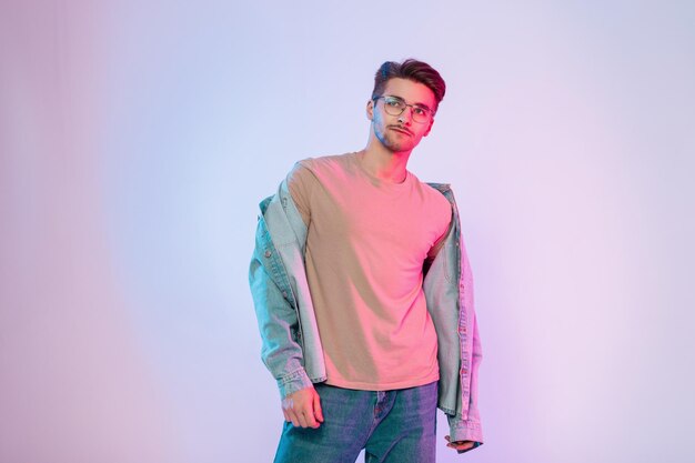 Photo stylish young handsome model hipster guy with hair and vintage glasses in a trendy beige tshirt and blue jeans shirt on a creative pink background in the studio