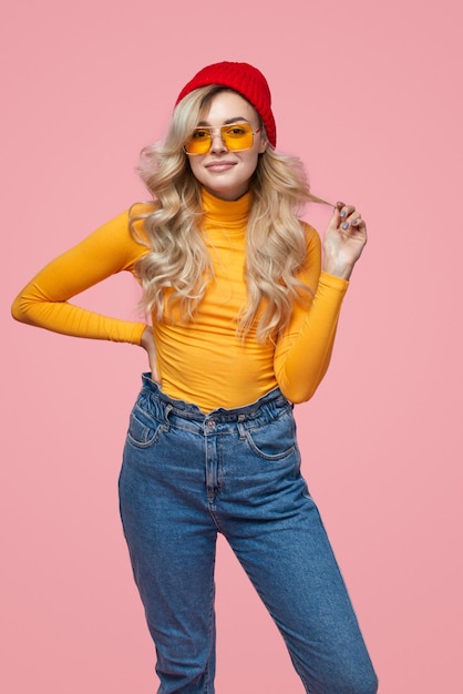 Stylish young lady touching hair and looking at camera in pink studio