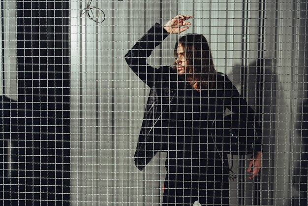 Stylish young long haired man in leather jacket holding helmet and looking through grid