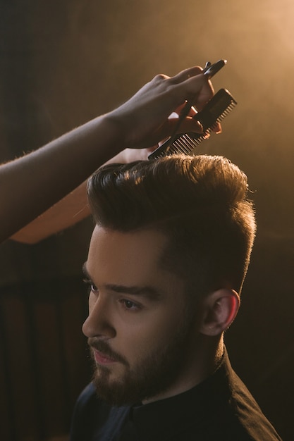 Stylish young male in hairdressing salon. Unrecognizable hairdresser cutting modern man hair, handsome guy portrait in focus on foreground, beauty concept