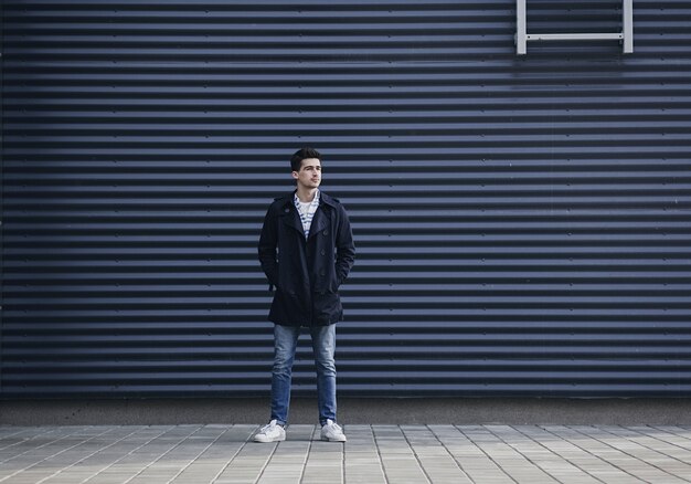 Stylish young man in a coat and jeans stands near the city wall