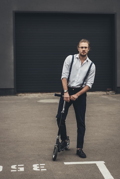 Stylish young man in eyeglasses and suspenders standing on scooter on parking