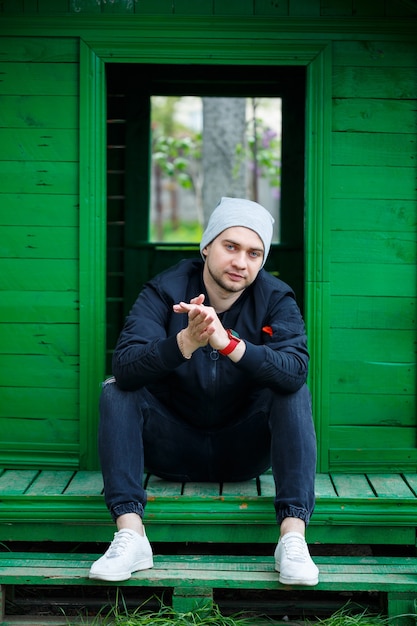 Stylish young man in a fashionable black jacket in blue jeans and hat posing on a sunny day near a green building outdoors. Modern guy on the street. Youth style