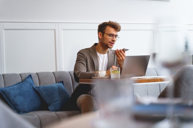 Stylish young man having phone conversation and smiling while sitting at the table with laptop in coffee house