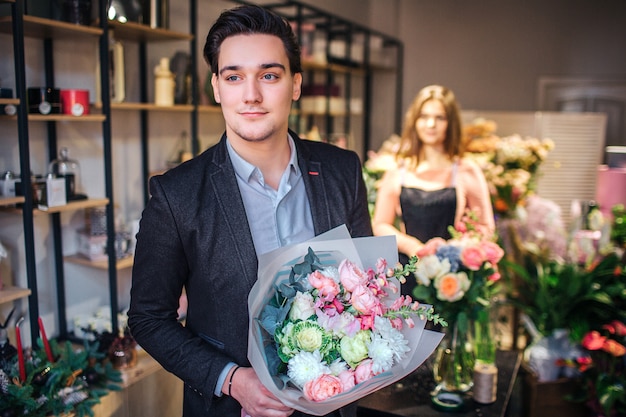 Stylish young man hold beautiful bouquet in hands and look forward. He goes out of flower shop. Young female florist stand behind him.