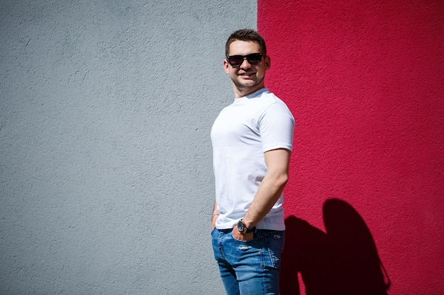 Стильный молодой человек, мужчина, одетый в белую пустую футболку, стоит на фоне серо-красной стены. Городской стиль в одежде, современный модный образ. Мужская мода