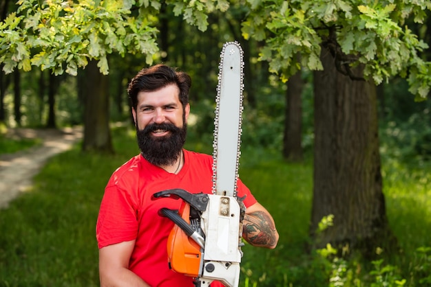 Stylish young man posing like lumberjack lumberjack worker standing in the forest with chainsaw defo...