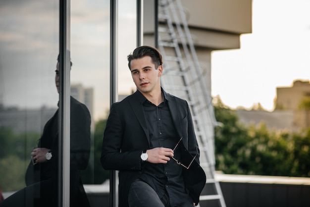 A stylish young man stands near a mirrored building during sunset. Fashion