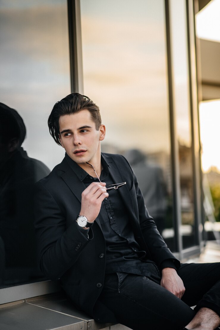 A stylish young man stands near a mirrored building during sunset. Fashion