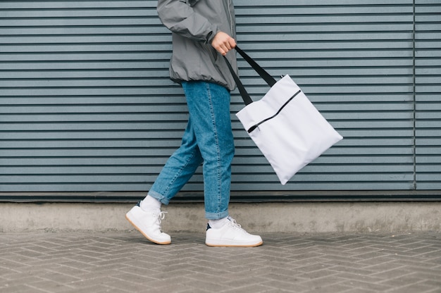 Giovane alla moda in abiti di strada a piedi con la borsa della spesa riutilizzabile su grigio