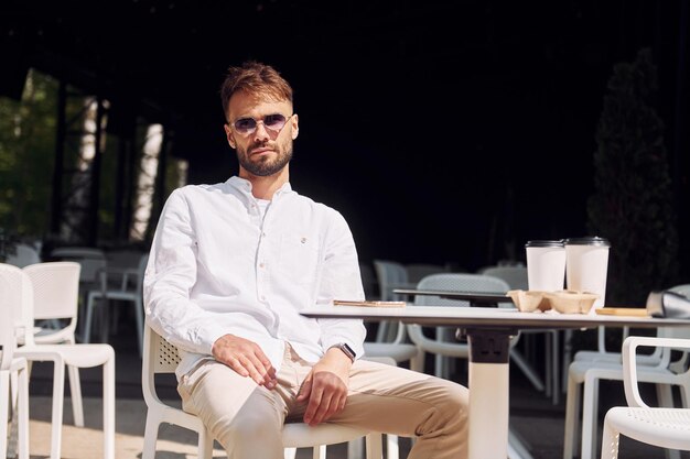 Stylish young man in sunglasses sitting on the chair of cafe outdoors