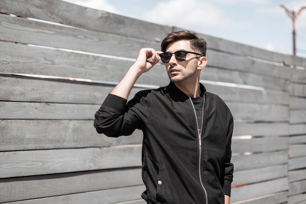 Stylish young man in a vintage black jacket in fashionable sunglasses with a trendy hairstyle relaxes near a wooden fence on a bright sunny day.American urban guy enjoys the sunshine outdoors.