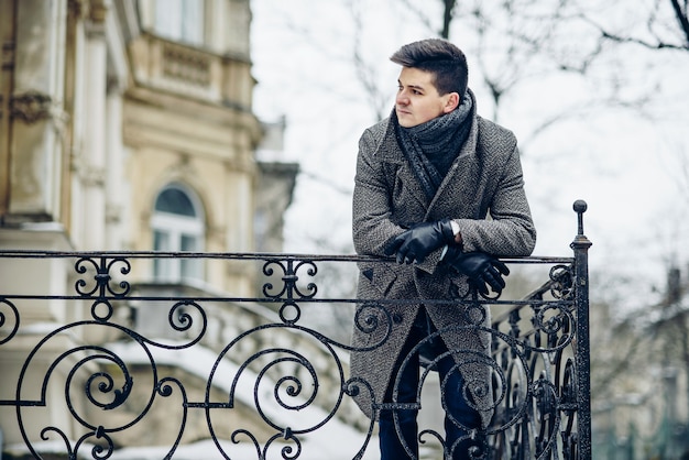 Un giovane uomo alla moda in un caldo cappotto grigio e guanti di pelle si appoggiò a un recinto forgiato