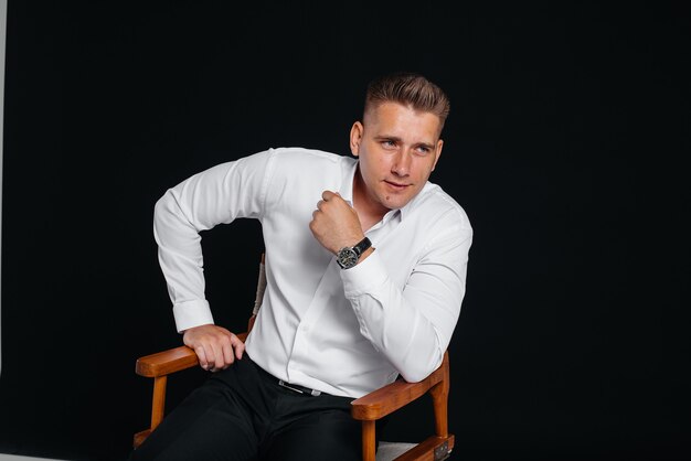 A stylish young man in a white shirt is sitting on the director's chair on a black background. A handsome charismatic businessman.