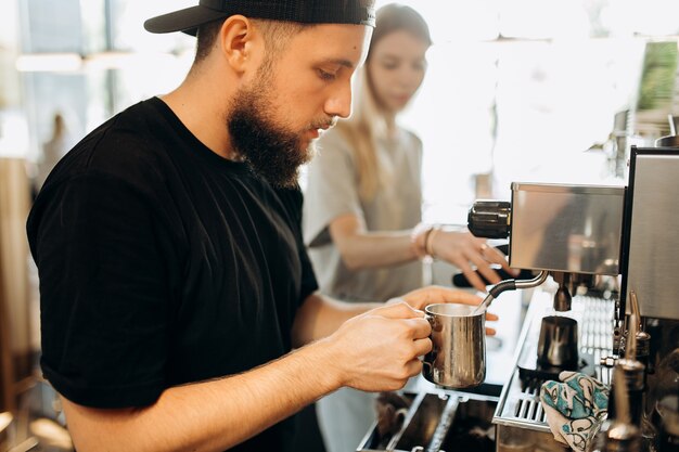 カジュアルな服を着て、ひげを生やしたスタイリッシュな若い男が、モダンなコーヒーショップのコーヒーマシンでコーヒーを調理します。 。