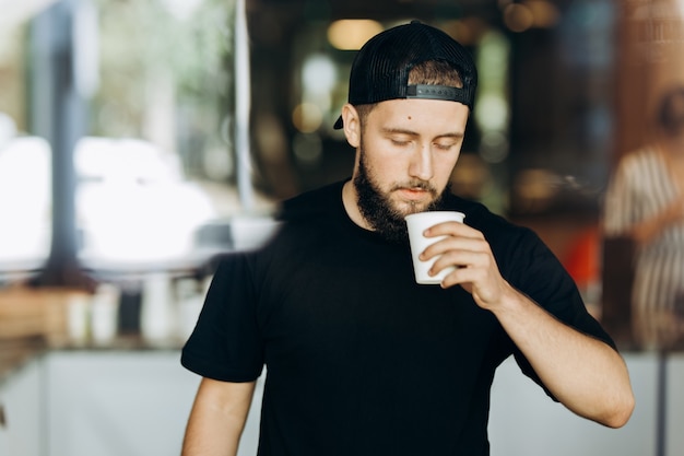 A stylish young man with beard,wearing casual outfit,stands and smells coffee in a modern coffee shop. .