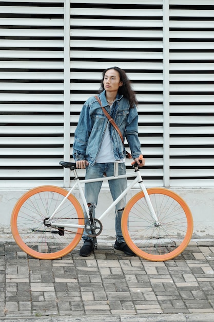 Stylish young man with long hair standing outdoors behind his bicycle and looking away