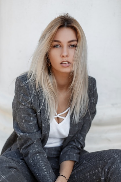 Stylish young model blond woman in a vintage gray suit sitting on the street near a white column