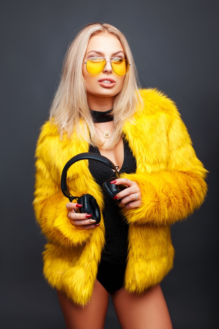 Photo stylish young model woman with headphones in yellow sunglasses and yellow fashion fur coat posing on gray