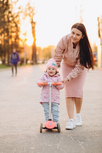 A stylish young mother in a pink clothes teaches a little daughter to ride a scooter.