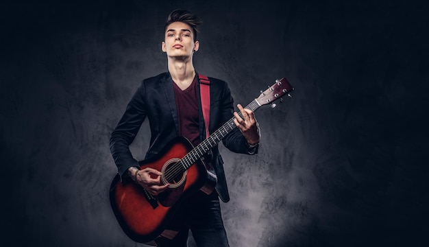 Giovane musicista alla moda con capelli alla moda in abiti eleganti con una chitarra in mano che suona e posa su uno sfondo scuro.