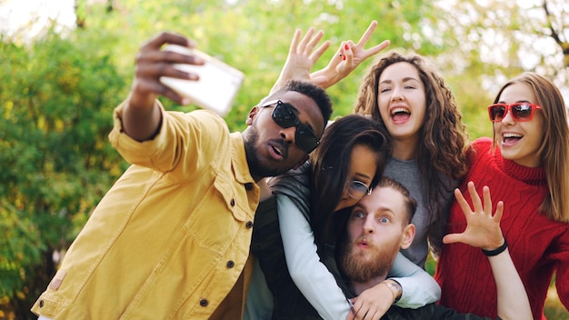 Stylish young people girls and guys are using smartphone to take selfie in park posing for camera and laughing Students are wearing casual clothing and sunglasses