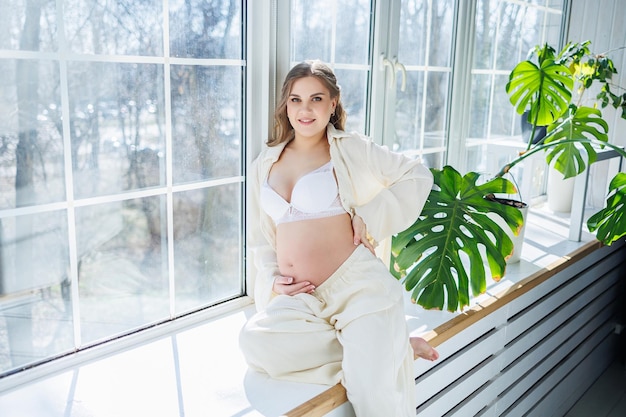 A stylish young pregnant woman in a white suit is sitting on the windowsill Pregnant woman in white underwear