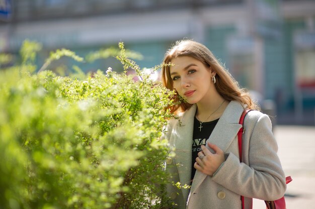 회색 코트를 입은 세련된 젊은 예쁜 소녀가 카메라를보고 도시 배경에 약간 미소를 짓습니다. 소녀는 녹색 덤불 옆에 서 있습니다.