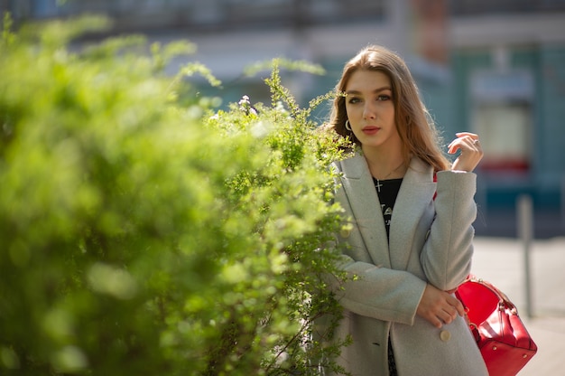 Stylish young pretty girl in a gray coat looks at the camera and smiles slightly against the background of the city. The girl stands by a green bush.