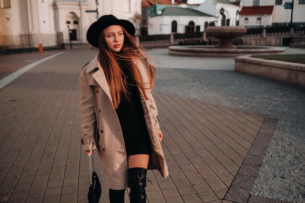 Elegante giovane donna in un cappotto beige in un cappello nero su una strada cittadina. moda di strada femminile. abbigliamento autunnale stile urbano.