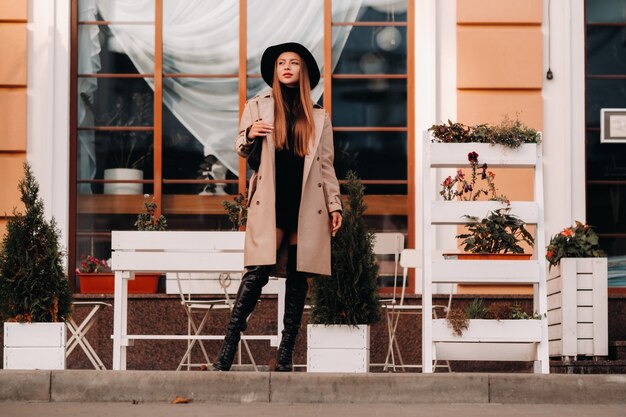 Photo stylish young woman in a beige coat in a black hat on a city street.