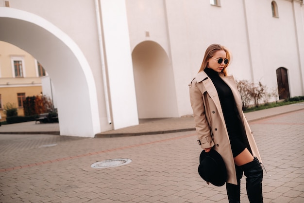 Stylish young woman in a beige coat and a black hat in his hands and glasses on a city street. Women's street fashion. Autumn clothing.Urban style.