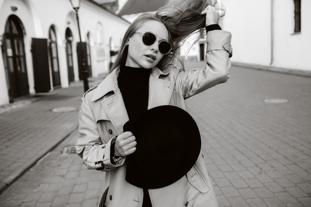 베이지 색 코트와 그의 손과 도시 거리에 안경에 검은 모자에 세련 된 젊은 여자. 여성의 스트리트 패션. 가을 의류. 도시 스타일.