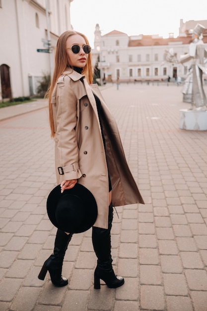 Elegante giovane donna in un cappotto beige e un cappello nero in mano e occhiali su una strada cittadina. moda di strada femminile. abbigliamento autunnale stile urbano.