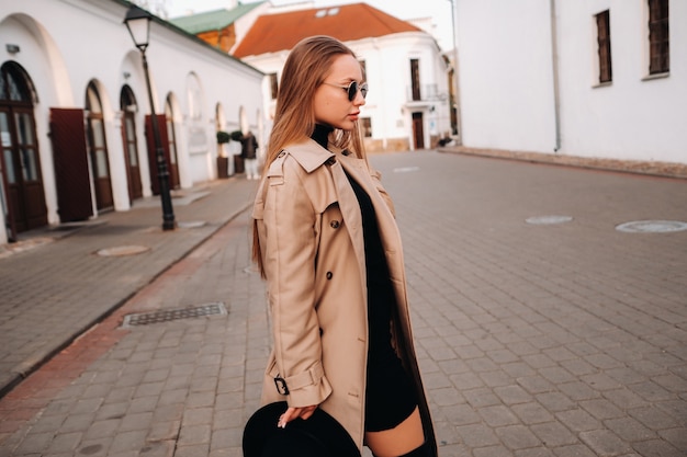 Stylish young woman in a beige coat and a black hat in his hands and glasses on a city street