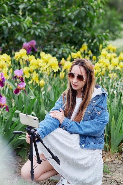 Stylish young woman blogger broadcasting a video using a tripod\
and phone among irises blogging lifestyle using modern\
technology