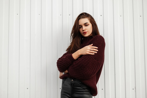 Stylish young woman in a burgundy sweater posing in the city near a white wall